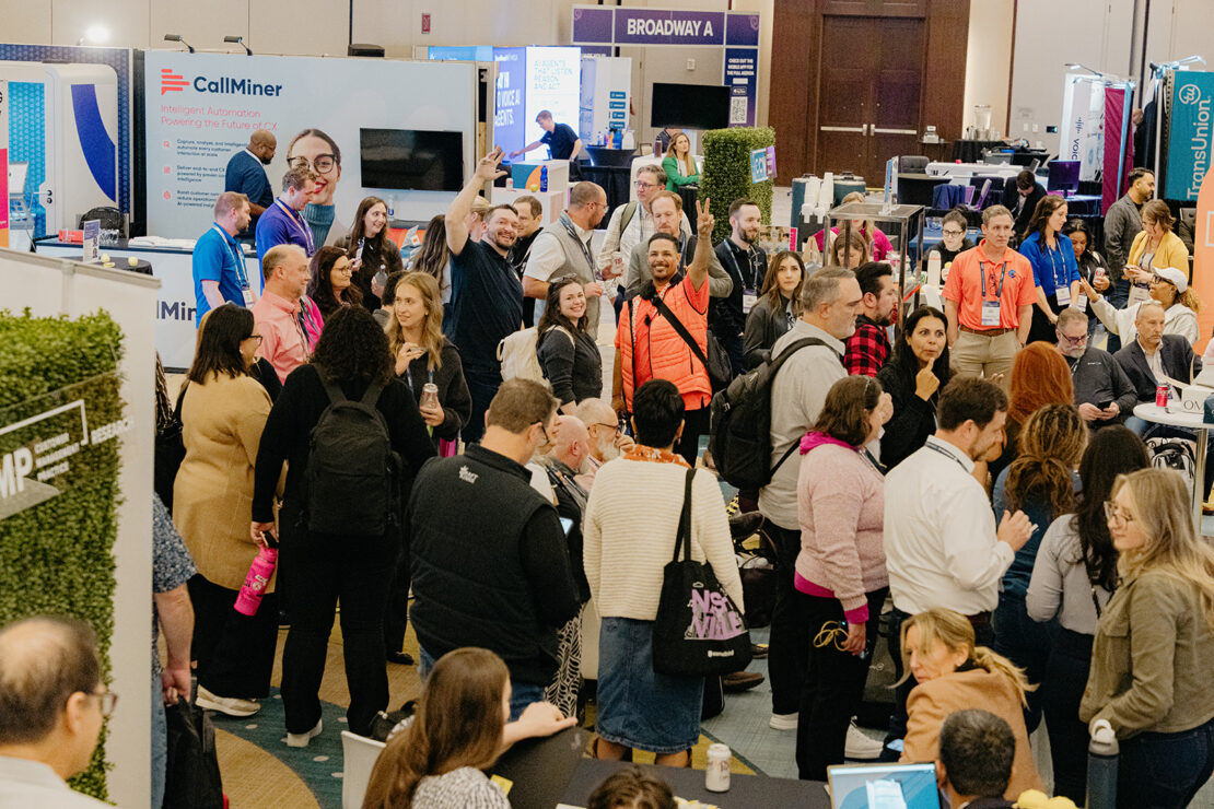 A large group of people attend a busy indoor business expo with booths, banners, and exhibitors engaging in conversation.