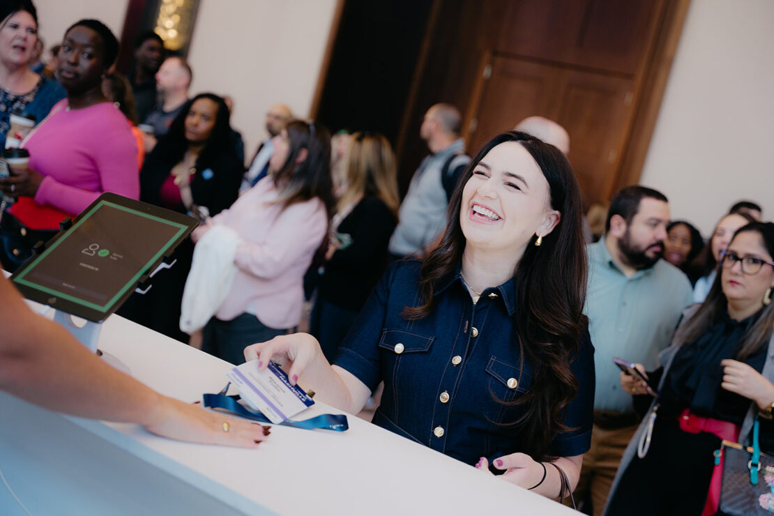 A woman smiles while handing her badge to a registration attendant at a counter, with a group of people waiting in line behind her.