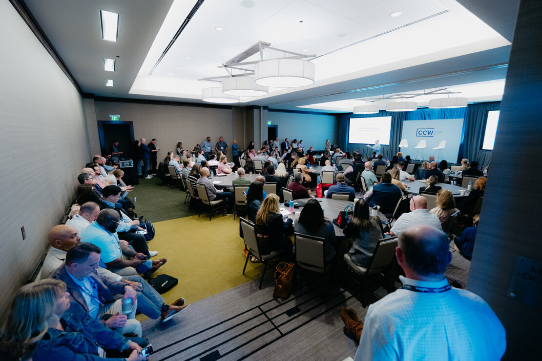 Conference room filled with attendees seated and standing, listening to speakers at the front near a CCW sign, with a projector screen and bright overhead lighting.