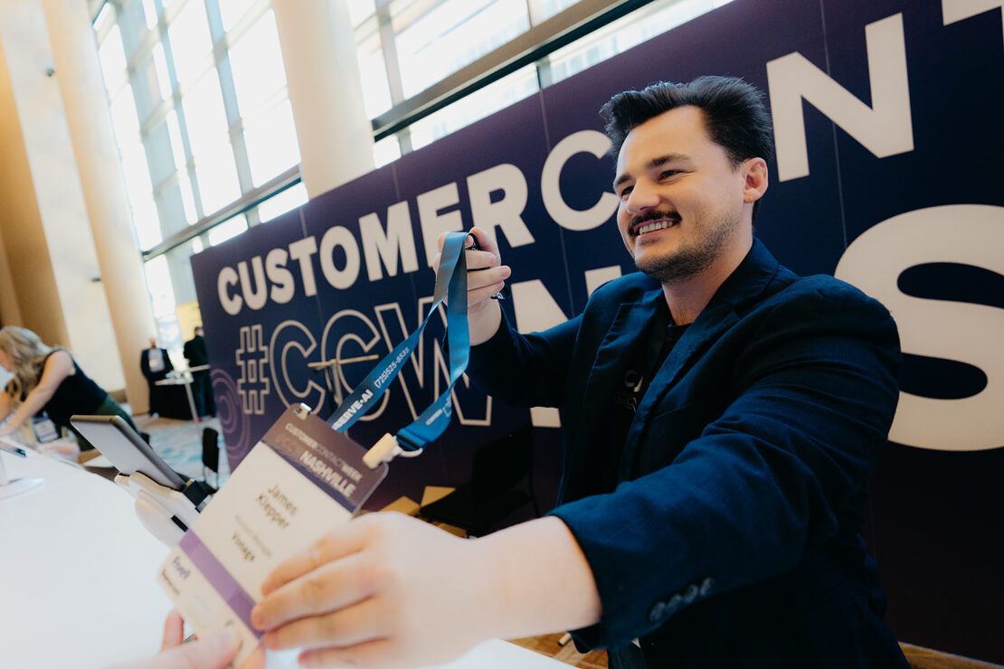 A man hands a conference badge to an attendee at a registration table in a brightly lit event space with "CUSTOMERCON" signage in the background.
