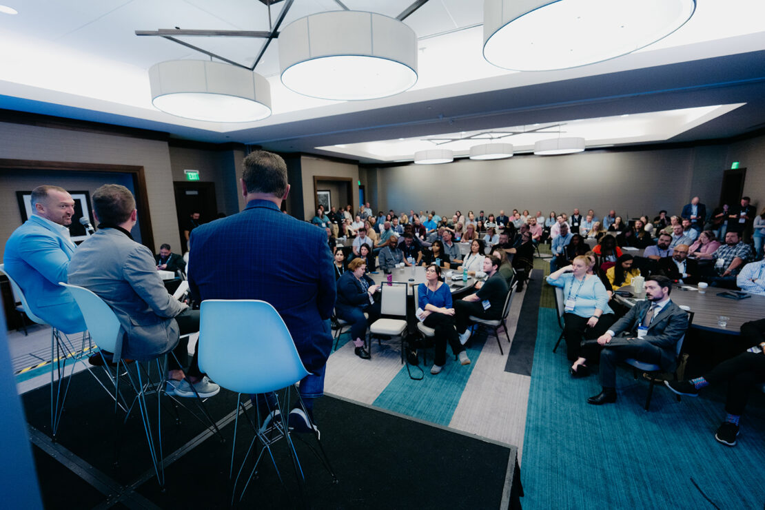 Four panelists sit on a stage facing an audience in a conference room, with people seated and listening attentively to the discussion.
