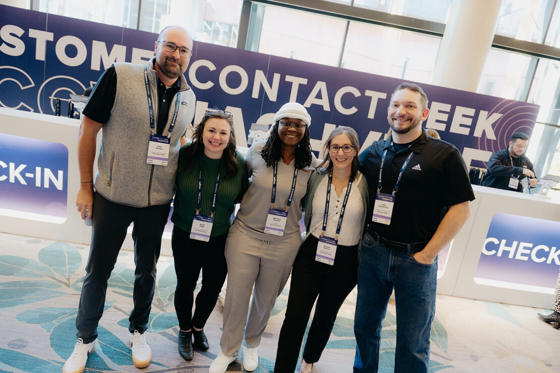 Five people standing and smiling in front of a check-in counter at an event, wearing name badges and casual business attire.