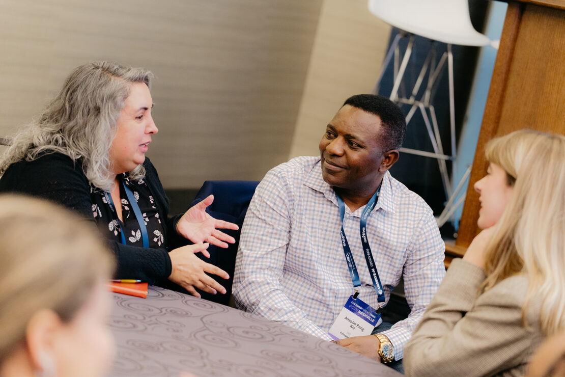 Three people sit around a table engaged in conversation at a professional event, wearing name badges and business attire.