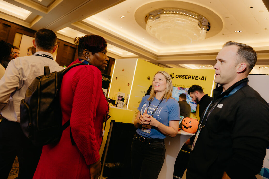 People talk and network at a conference booth with "OBSERVE.AI" signage; one person holds a pumpkin bucket under bright ceiling lights.