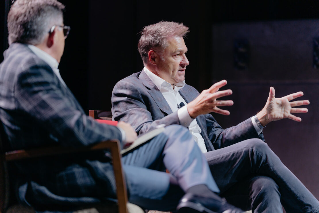 Two men in suits sit and talk on stage; one gestures with his hands while speaking, and the other listens with a notebook in his lap.