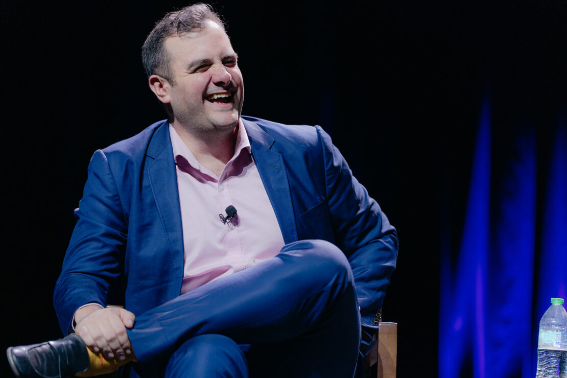 A man in a blue suit and pink shirt sits on a chair, smiling and laughing, with a microphone clipped to his shirt and a water bottle beside him.