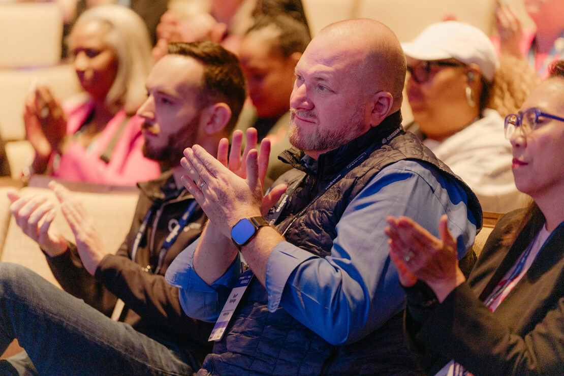 A group of people sit in an audience clapping, with focus on a man in a blue shirt and black vest in the center.