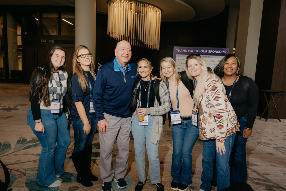 A group of seven people, including six women and one man, stand closely together smiling for a photo at an indoor event with sponsor signage in the background.
