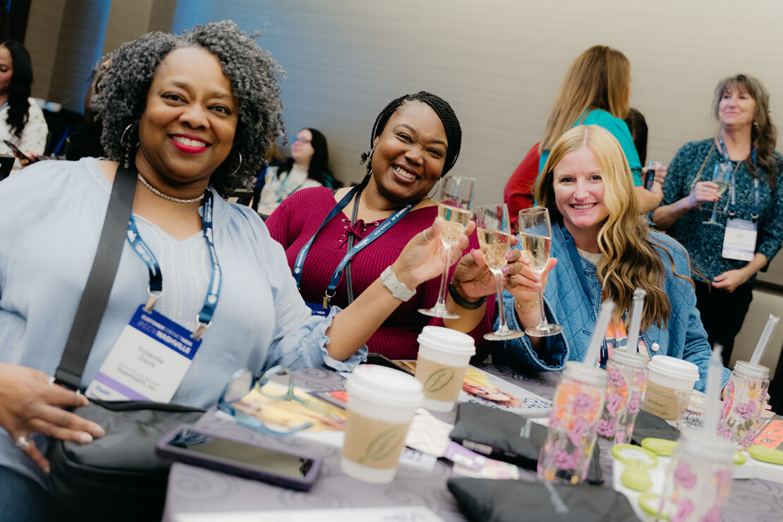 Three women sit at a table, smiling and holding up glasses in a toast at a conference or event, with coffee cups and event badges visible.