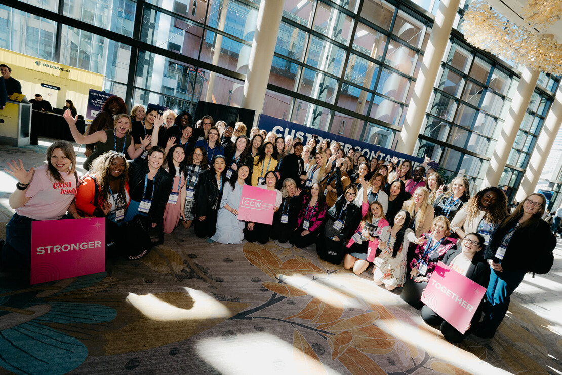 A large, diverse group of people pose together indoors at an event, holding signs that read “STRONGER” and “TOGETHER” in front of large windows.