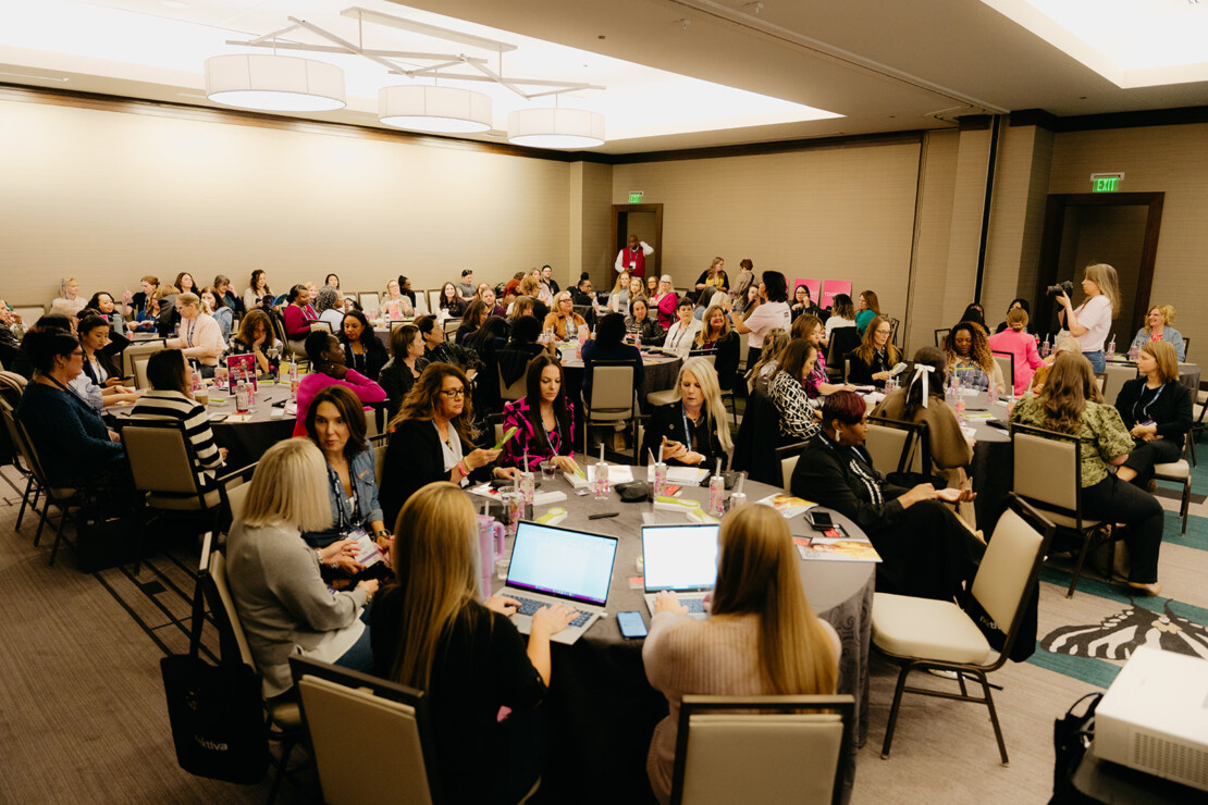 A large group of people sit at round tables in a conference room, some using laptops, while a woman stands and speaks at the front.