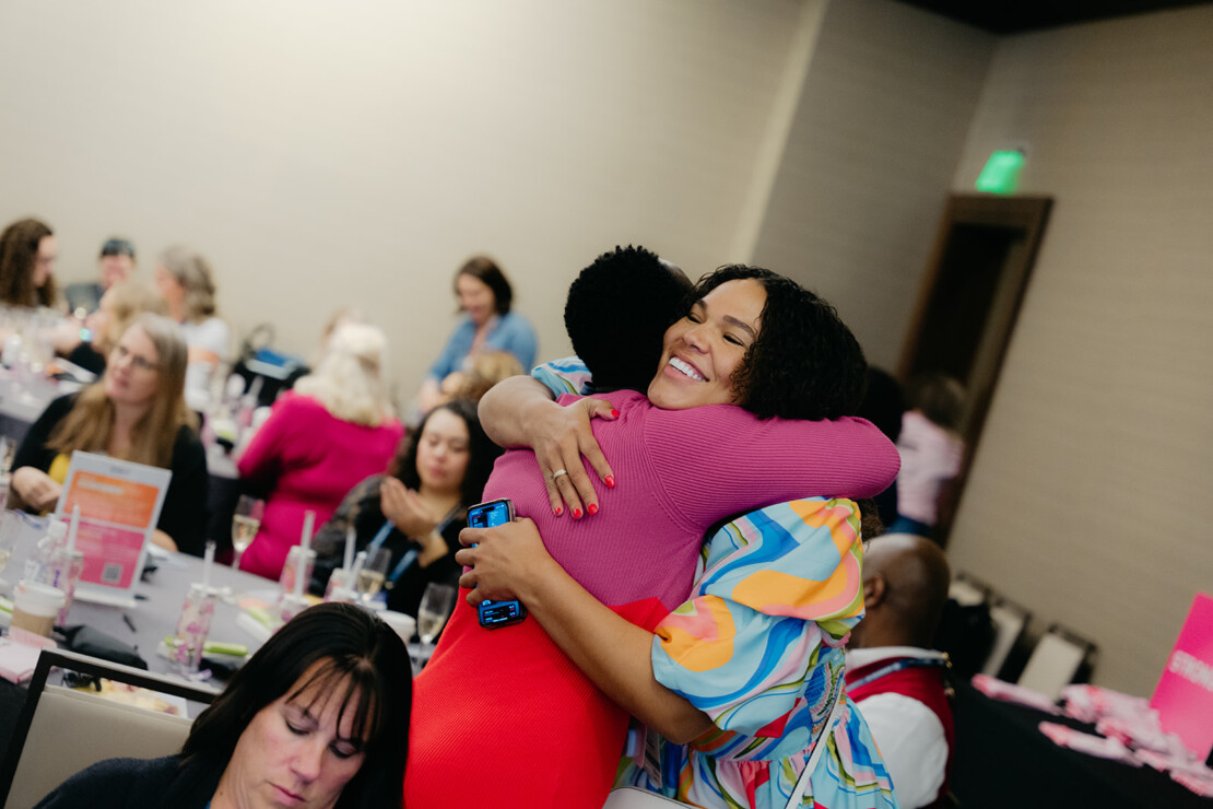 Two women hug and smile at a crowded indoor event, while other attendees sit at tables in the background.