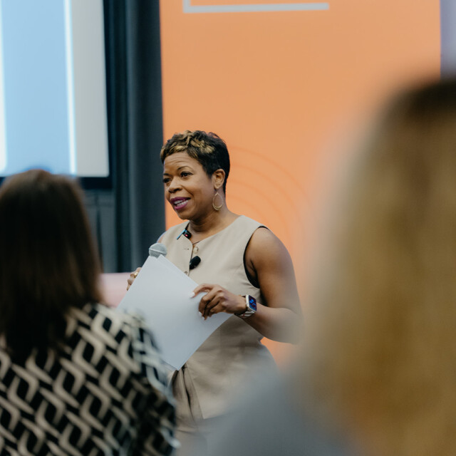 A woman stands speaking with a microphone and holding papers in front of an orange “CCW Customer Contact Week” sign, addressing an audience.
