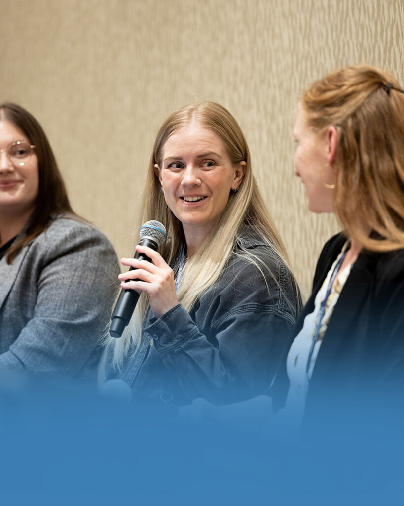 Three women sit side by side indoors; the woman in the center holds a microphone and looks at the woman on the right while speaking.
