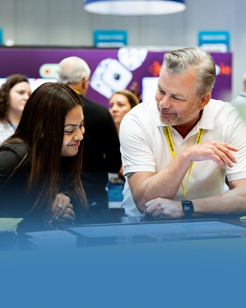 Two people sit at a table, looking at documents and talking, with a busy expo or conference environment in the background.