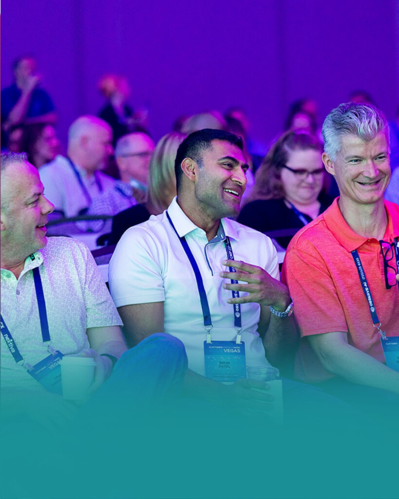 Three men sitting among a larger audience at an event, talking and smiling, all wearing lanyards and casually dressed.