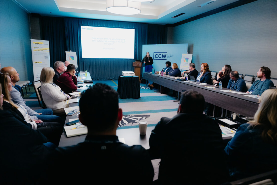 A group of people sit around a U-shaped table, listening to a presenter speaking at the front of a conference room with a screen and CCW banners.