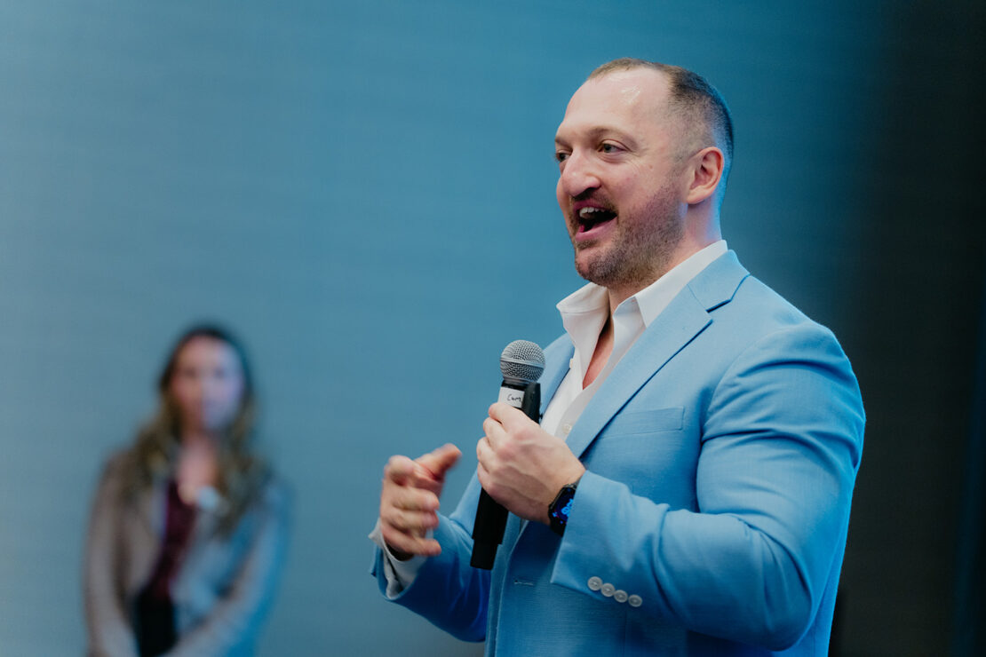 A man in a light blue suit speaks into a microphone, gesturing with his hands, while a woman stands in the blurred background.