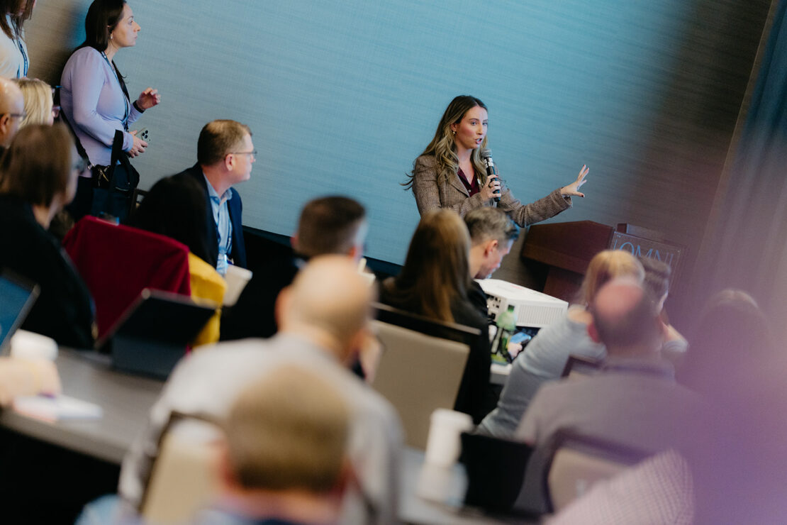 A woman stands at a podium speaking into a microphone to an audience seated at tables during a conference or seminar.