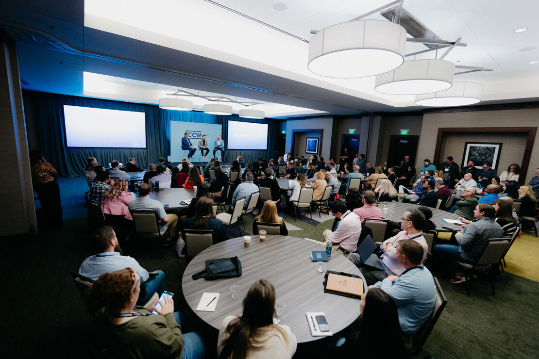 A large group of people seated in a conference room listens to a presentation with slides displayed on twin screens at the front of the room.