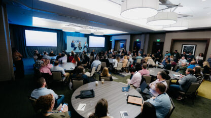 A large group of people seated in a conference room listens to a presentation with slides displayed on twin screens at the front of the room.