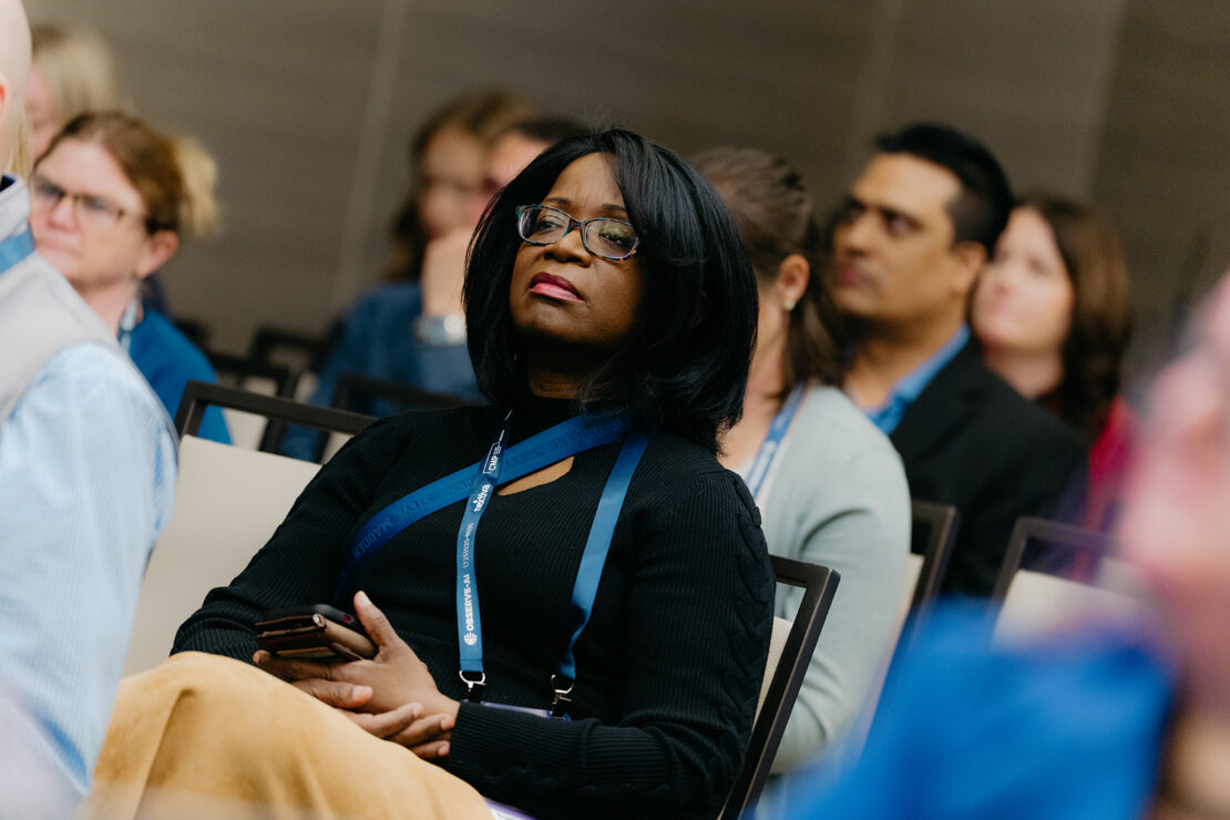 A woman holding a phone sits attentively among a group of people at an indoor event or conference.