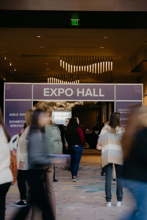People walk through the entrance of an expo hall with a large sign overhead. The hallway is well-lit with modern chandeliers.