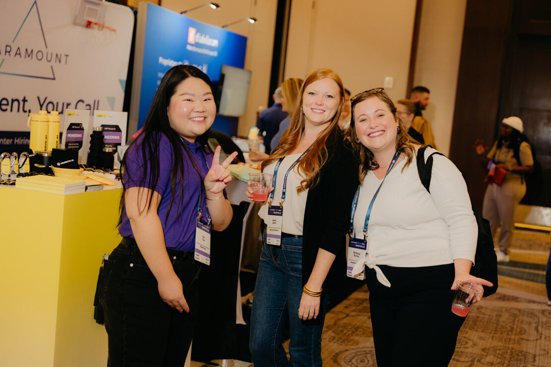 Three women pose and smile at a conference or expo, each wearing name badges and holding drinks, with display booths and attendees visible in the background.