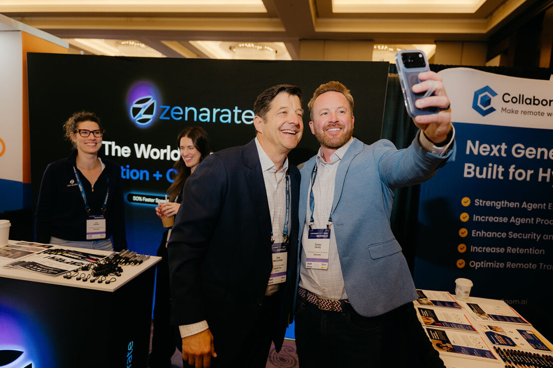Two men in business attire take a selfie at a conference booth, with two women standing nearby and promotional banners in the background.