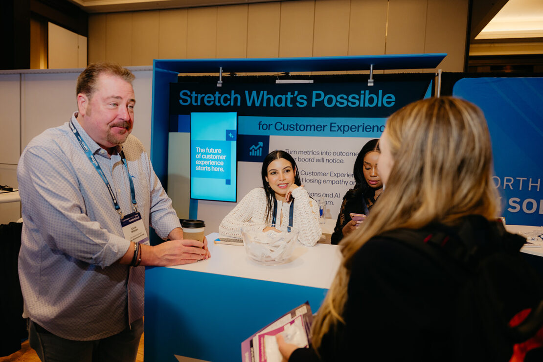 A man talks to three women at a booth with a sign reading "Stretch What's Possible for Customer Experience" at a conference or event.