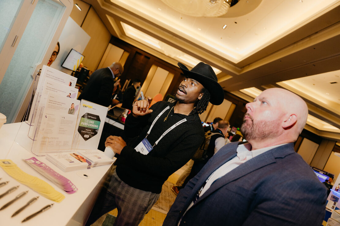Two men stand at a conference booth, one gesturing while speaking, with brochures and promotional materials visible on the table.