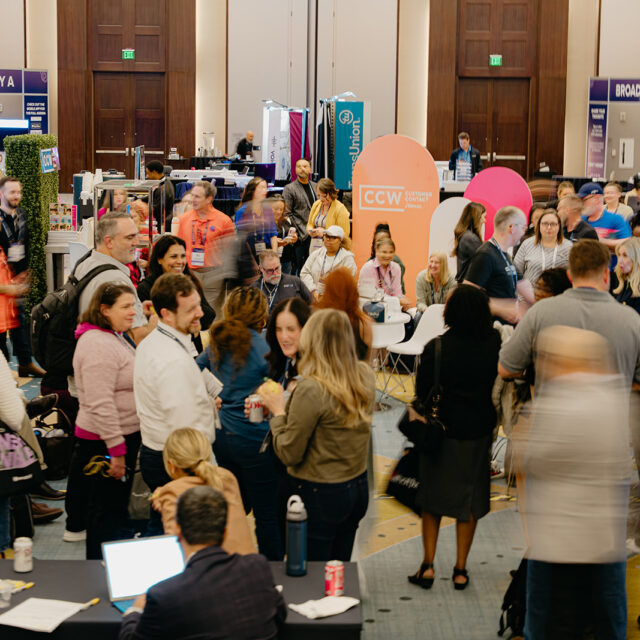 A large group of people gathers and interacts at a busy indoor conference or expo, with booths and banners visible in the background.