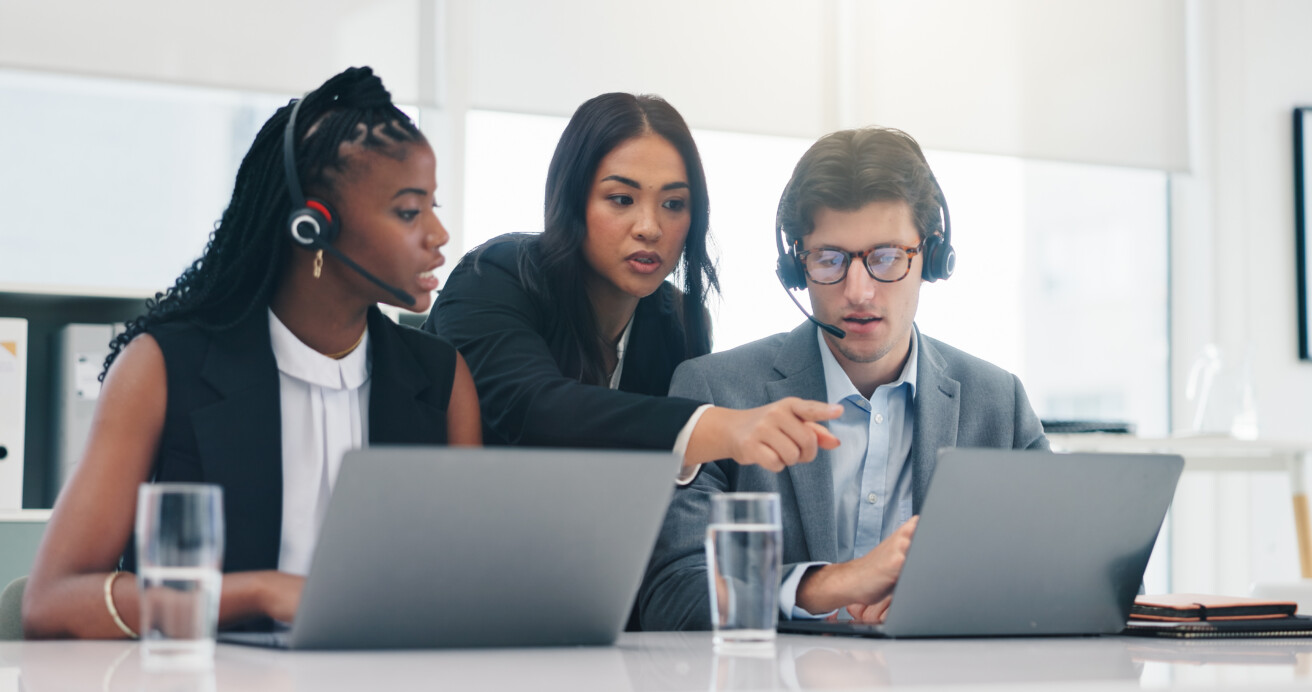 Three coworkers with headsets sit at a desk with laptops and glasses of water. One person stands and points at a colleague’s screen, suggesting collaboration or technical support.