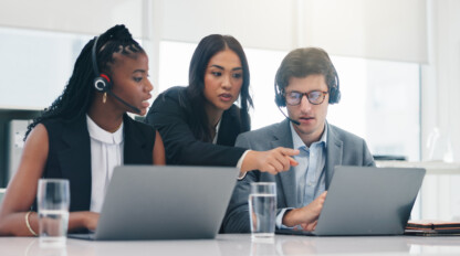 Three coworkers with headsets sit at a desk with laptops and glasses of water. One person stands and points at a colleague’s screen, suggesting collaboration or technical support.