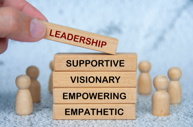 A hand places a wooden block labeled "LEADERSHIP" on top of blocks labeled "SUPPORTIVE," "VISIONARY," "EMPOWERING," and "EMPATHETIC," surrounded by wooden figures.