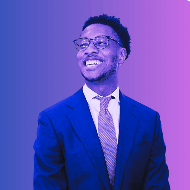 A man in a suit and tie smiles while looking to the side, standing against a blue and purple gradient background.