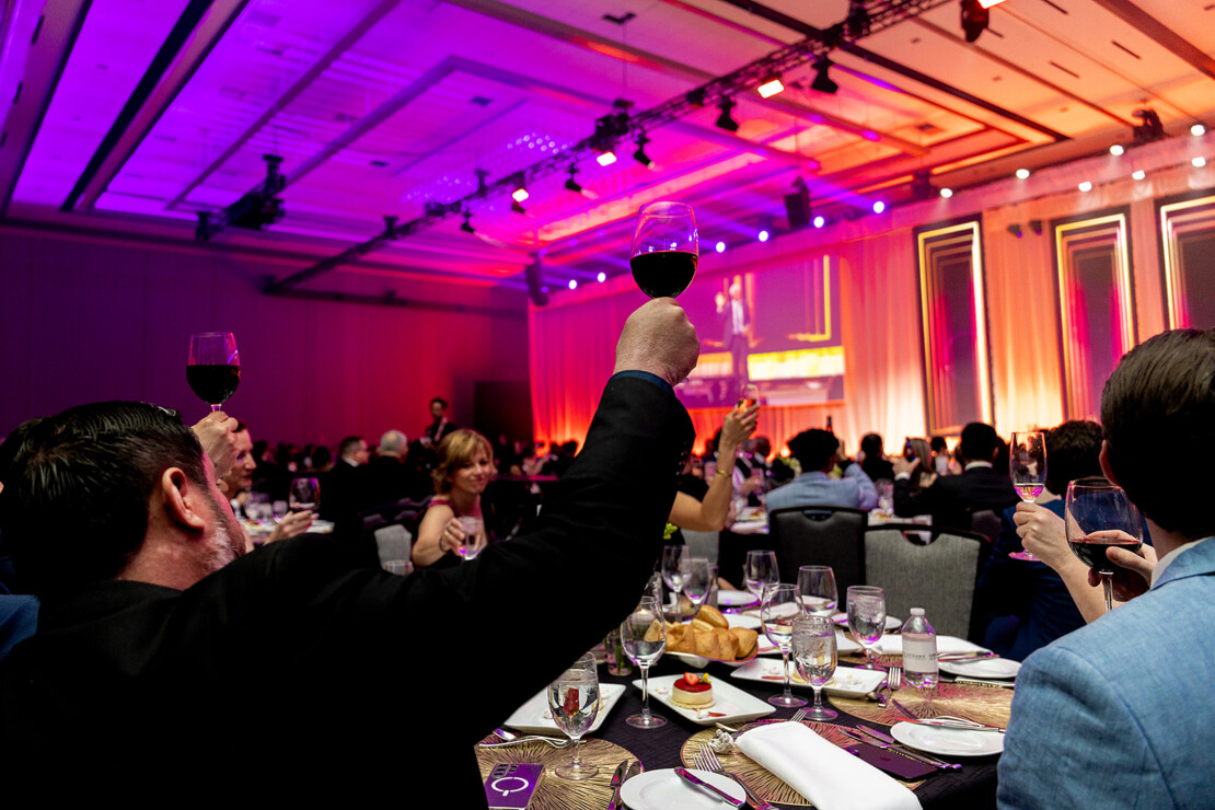 People seated at round tables raise wine glasses in a toast during a formal event in a large, brightly lit banquet hall.
