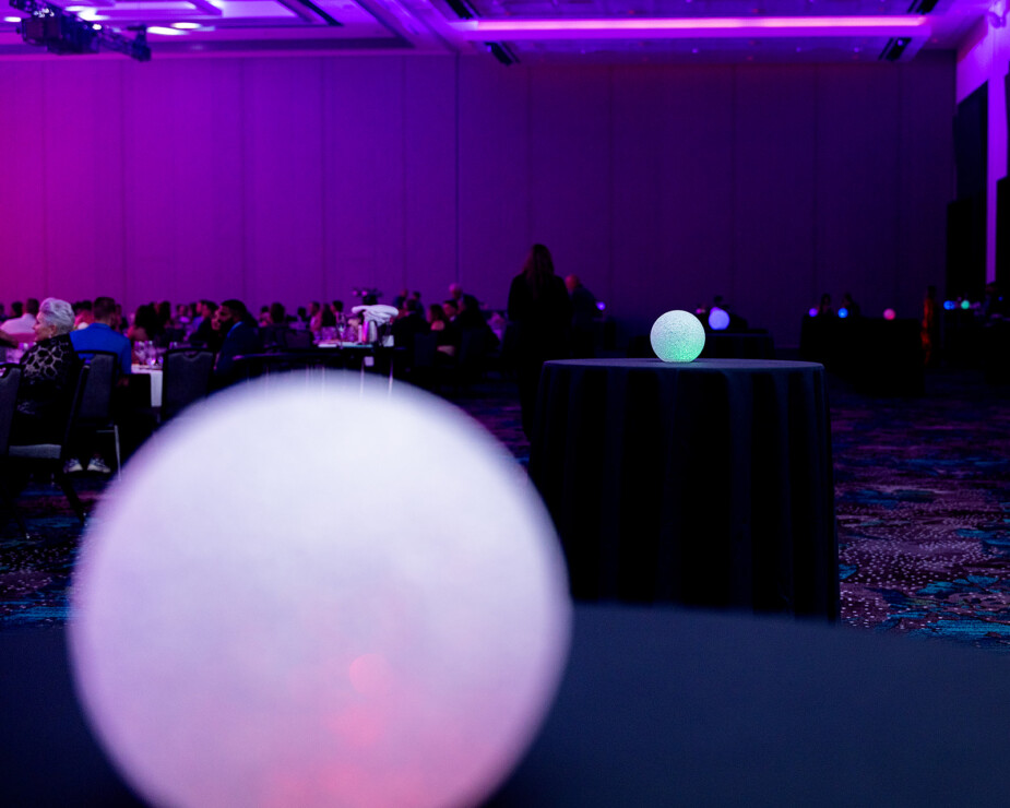 A round white light sits on a table in the foreground, with another similar light on a distant table in a dimly lit, purple event room filled with seated guests.