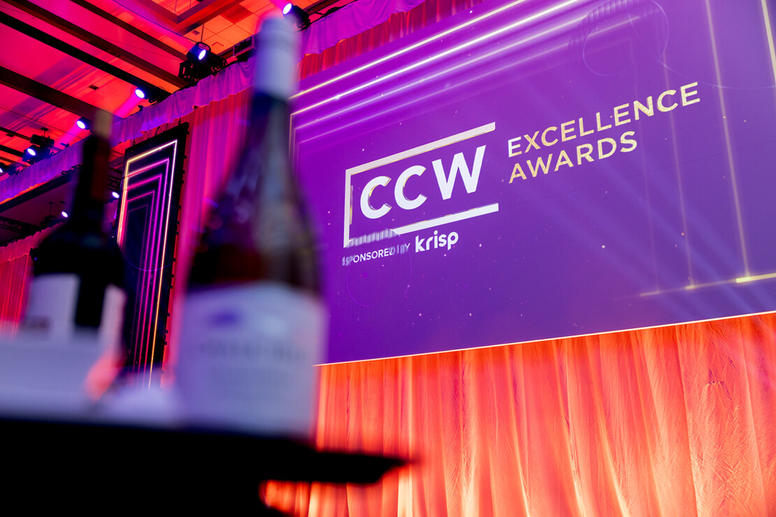 A large screen displays "CCW Excellence Awards" at an event, with bottles blurred in the foreground and orange stage curtains in the background.