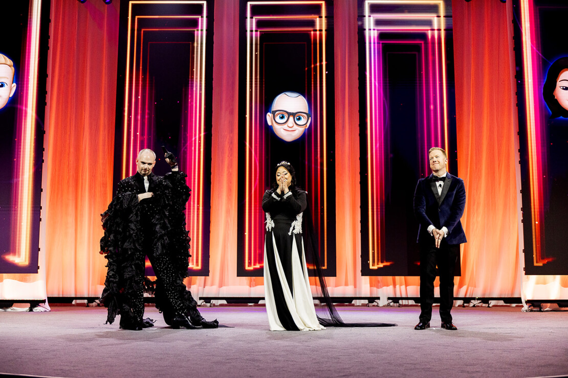 Three people in formal and dramatic attire stand on a stage with vibrant orange lighting and large digital avatars displayed on screens behind them.