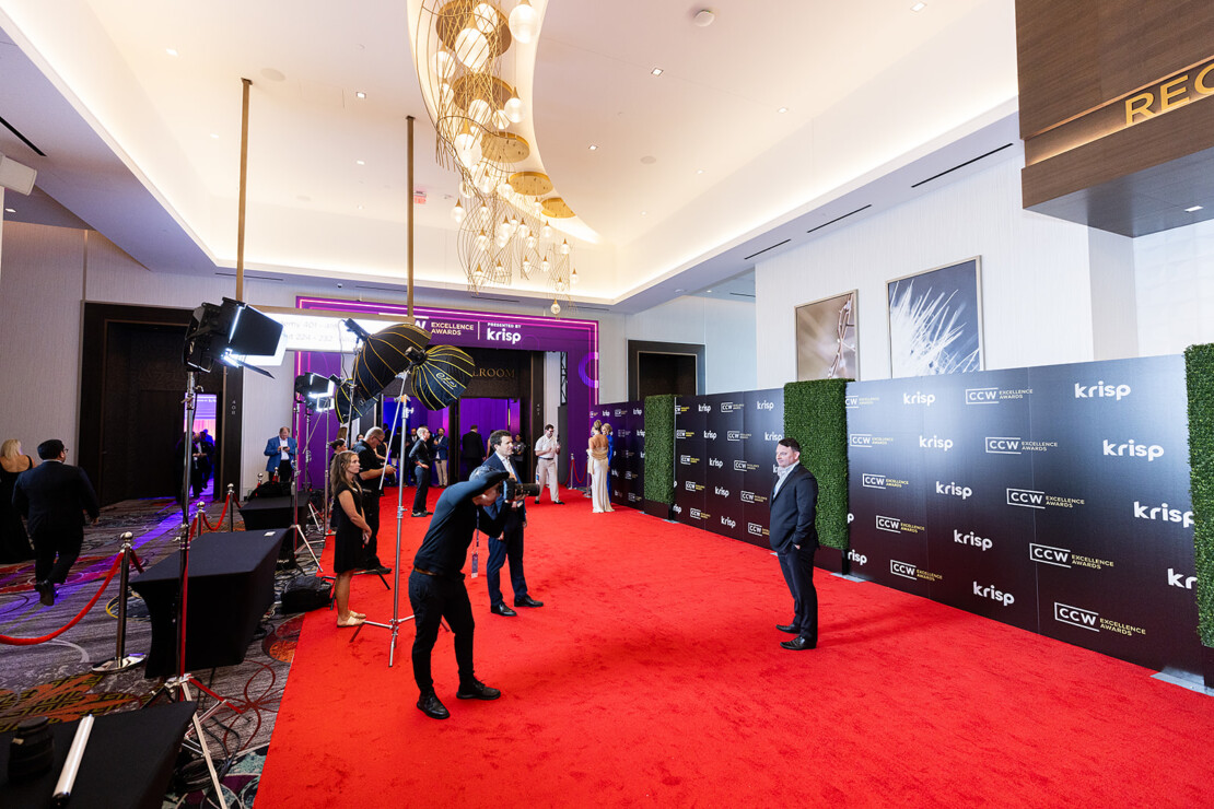 A man poses for photos on a red carpet at an event, with photographers, lighting equipment, and branded backdrop in a modern, well-lit lobby.