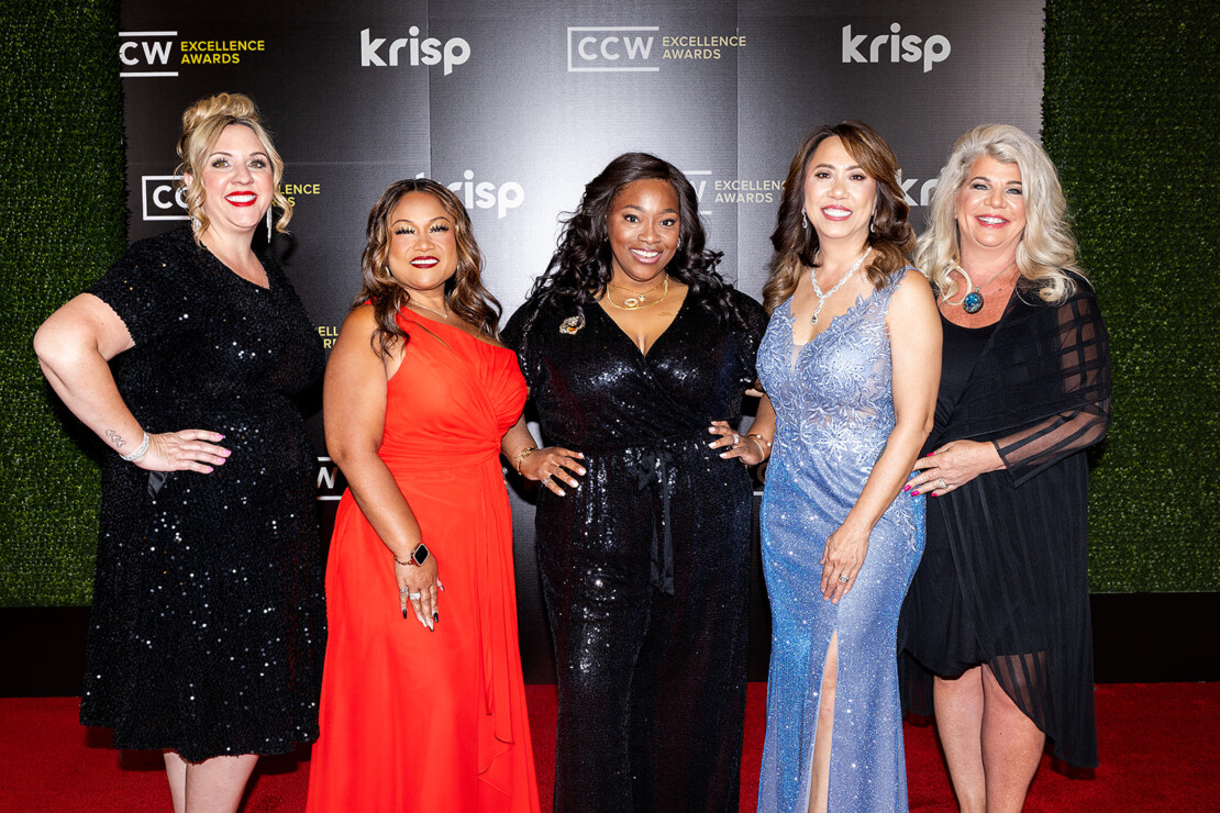 Five women in formal dresses pose together on a red carpet at an event with a backdrop featuring logos for CCW Excellence Awards and Krisp.