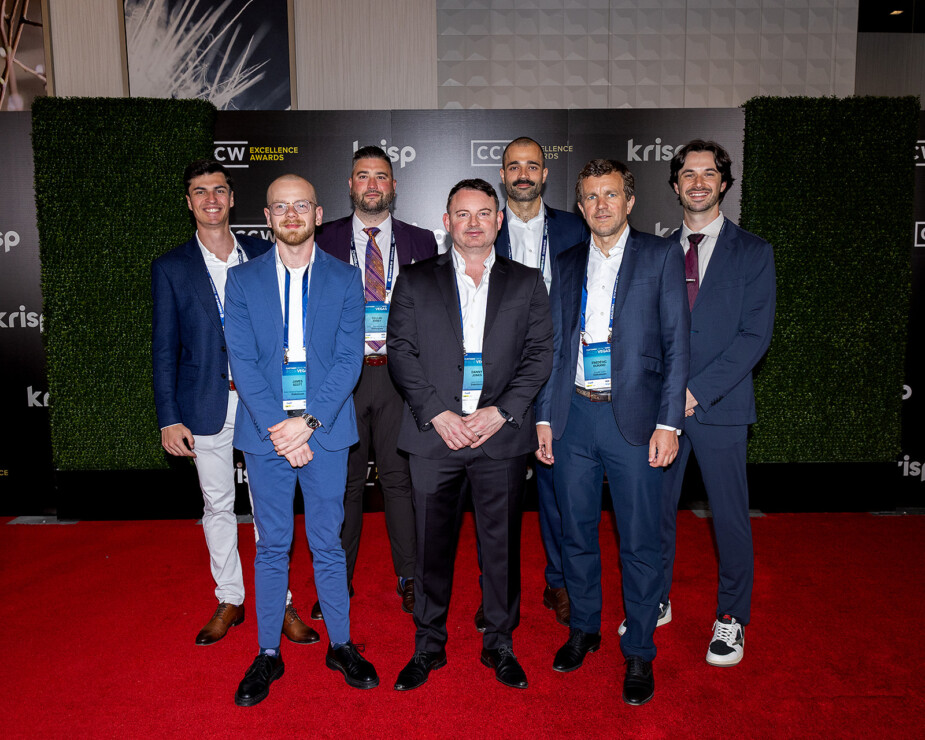 Seven men in business attire stand on a red carpet in front of a green wall backdrop with event signage and logos visible.