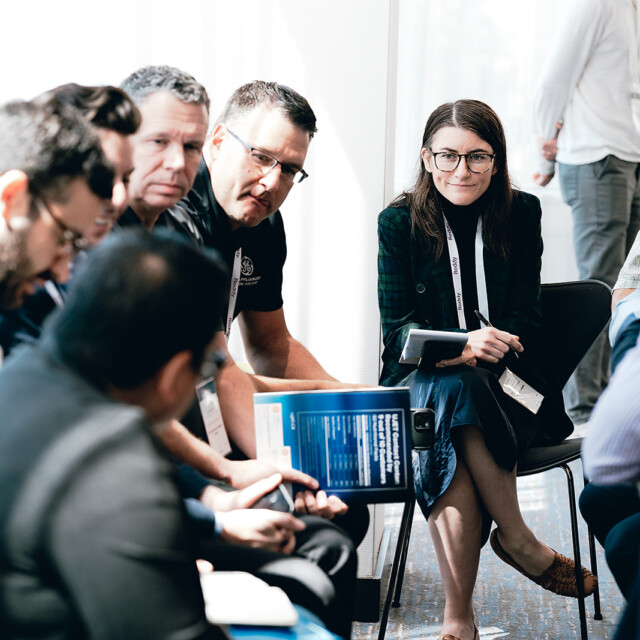 A group of people sit in a circle having a discussion; one woman with glasses faces the camera, holding a notebook.