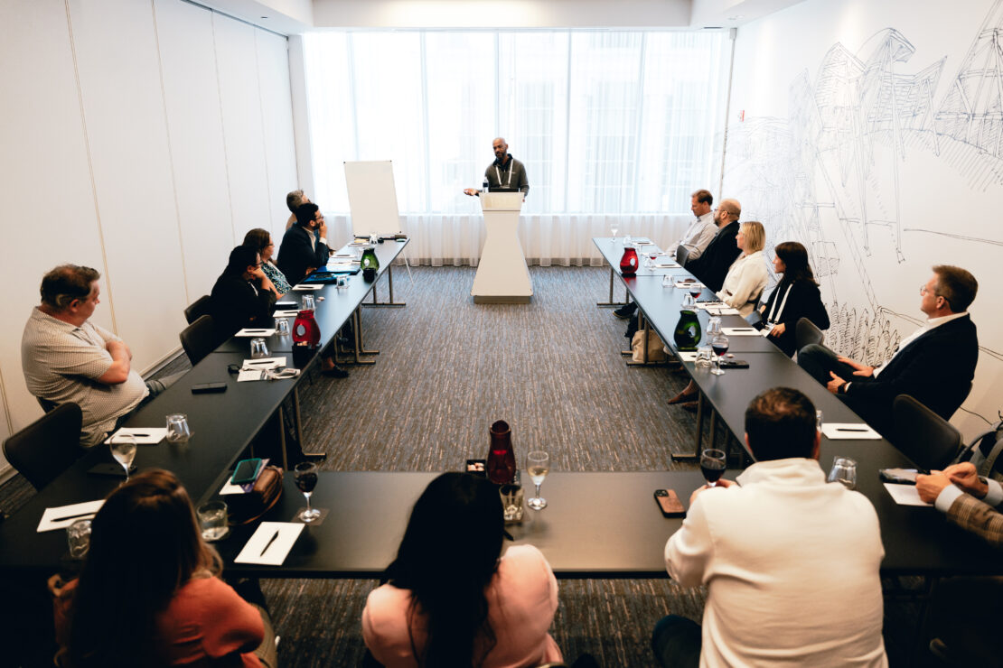 A group of people sits around a U-shaped table in a conference room, listening to a speaker standing at a podium in front of large windows.