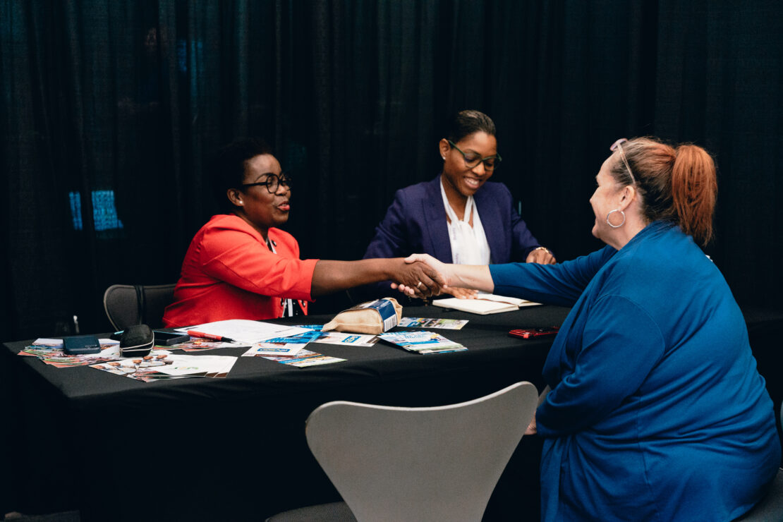 Two women sit at a table across from a third woman, shaking hands and smiling during a professional meeting or interview. Papers and brochures are spread across the table.