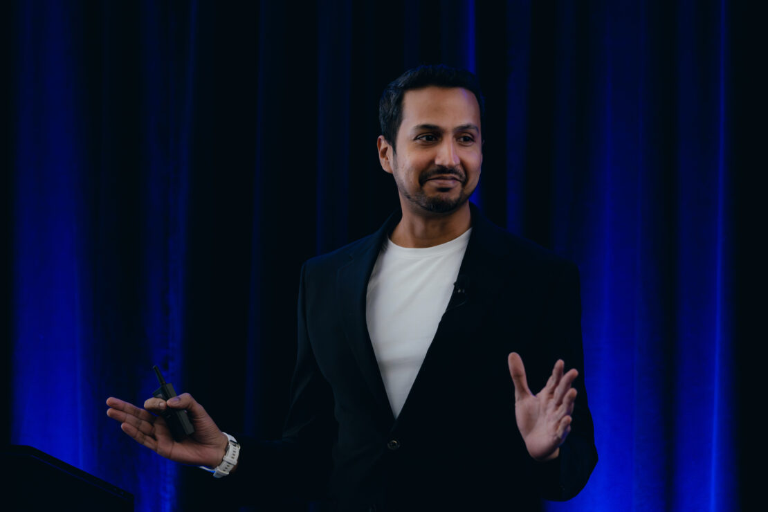 A man in a black blazer and white shirt gestures with his hands while speaking on stage in front of dark blue curtains.