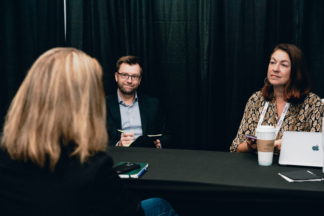 Three people sit at a table in front of a black curtain; two interviewers face the camera with notebooks and laptops, while the interviewee’s back is visible in the foreground.