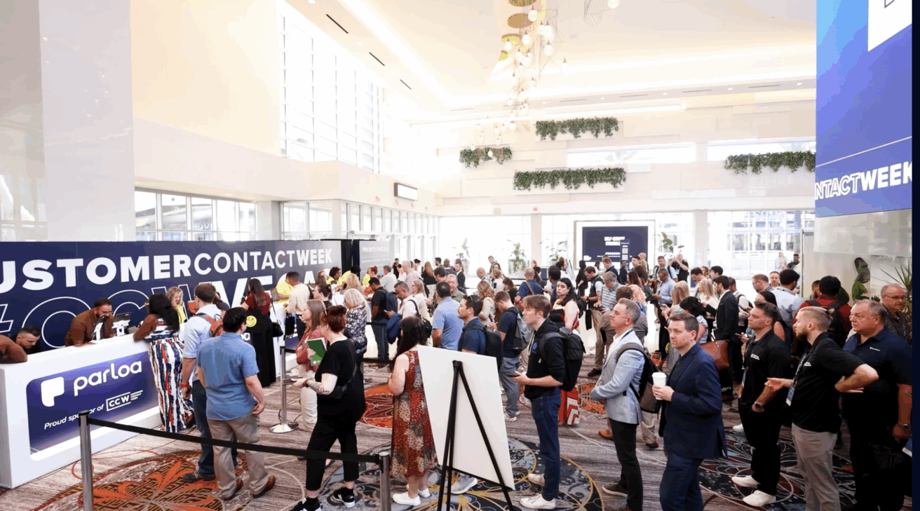 A large group of people stands in line and gathers in a well-lit lobby at a Customer Contact Week event, with banners and registration booths visible.