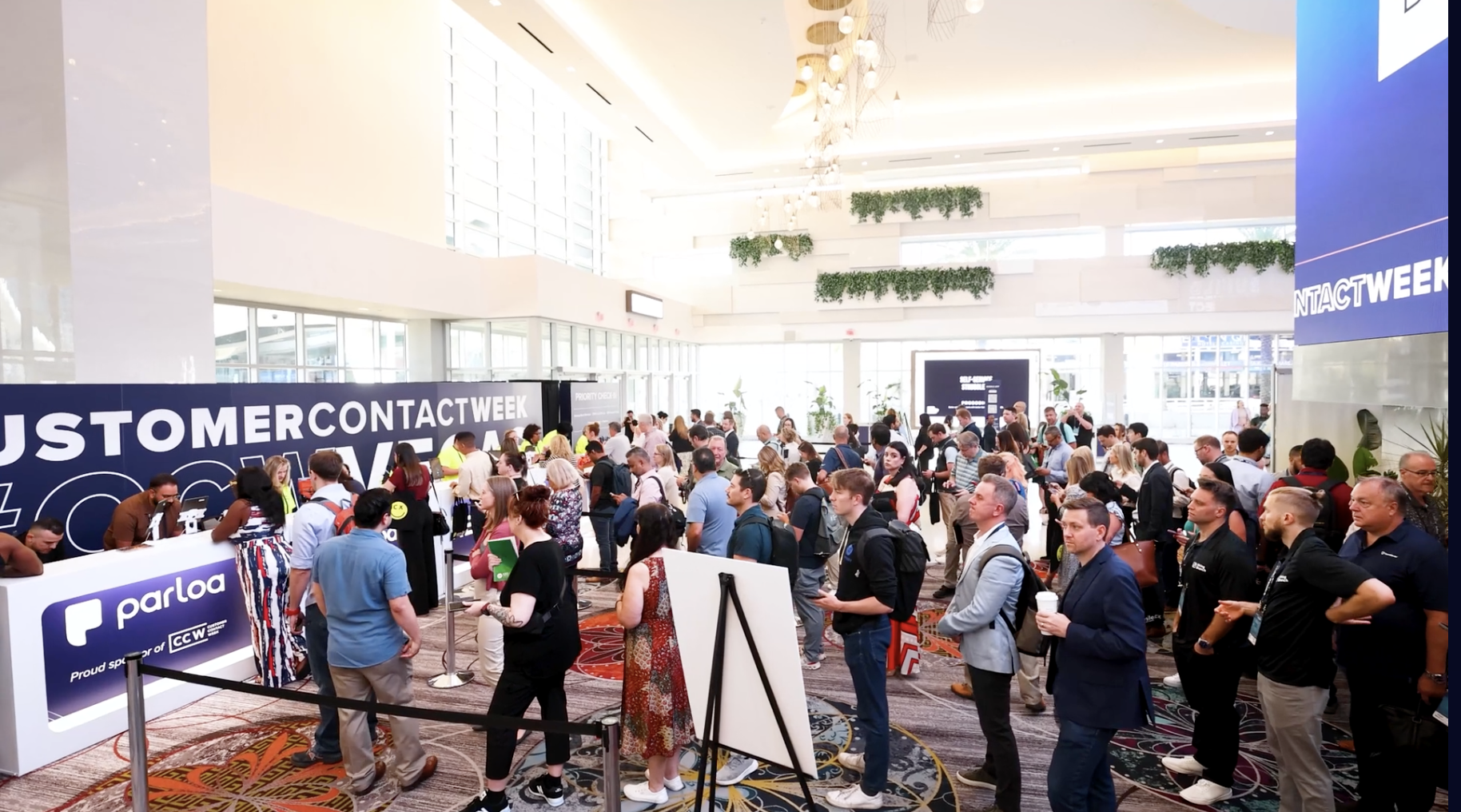 A large group of people stands in line and gathers in a well-lit lobby at a Customer Contact Week event, with banners and registration booths visible.